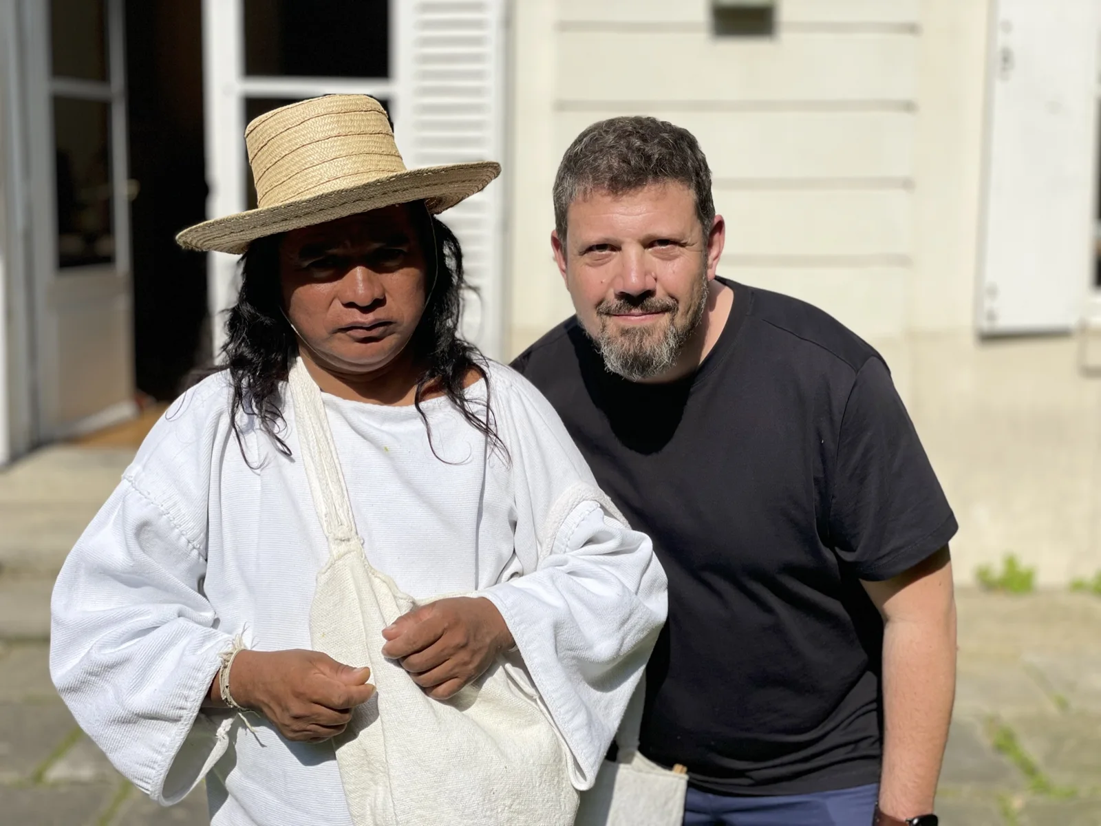Mihai Dragomirescu standing beside a Kogi man dressed in white traditional clothing, a woven straw hat and a woven shoulder bag, in front of a pale building in the sun