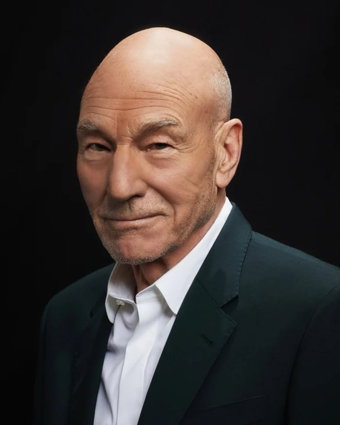 Sir Patrick Stewart, smiling a little, in a dark jacket and a white shirt against a black background.