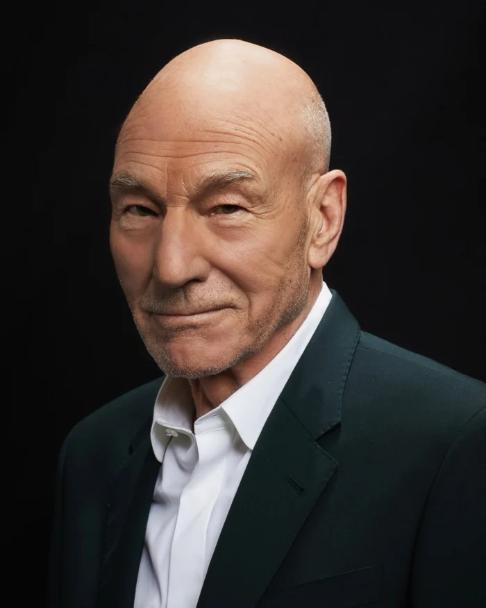 Sir Patrick Stewart, smiling a little, in a dark jacket and a white shirt against a black background.
